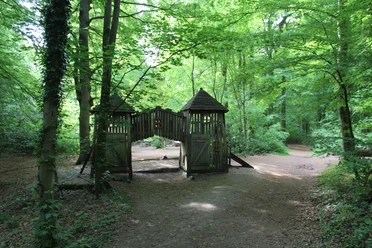 Ein naturnaher Spielplatz im Wald mit einer Holzburg und umgebenden hohen Bäumen unter grünem Blätterdach.