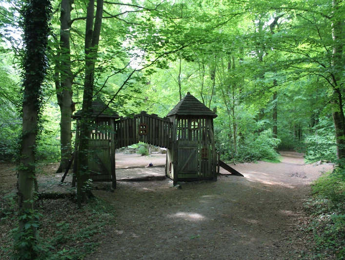 Ein naturnaher Spielplatz im Wald mit einer Holzburg und umgebenden hohen Bäumen unter grünem Blätterdach.
