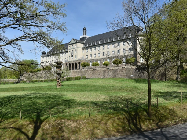 Thomas-Morus-Akademie Das Bild zeigt ein historisches Gebäude auf einer Anhöhe, umgeben von grünen Bäumen und Rasenflächen.