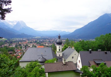 St. Anton Blick über Garmisch-Partenkirchen mit Zwiebelturm und umliegenden Bergen unter wolkigem Himmel