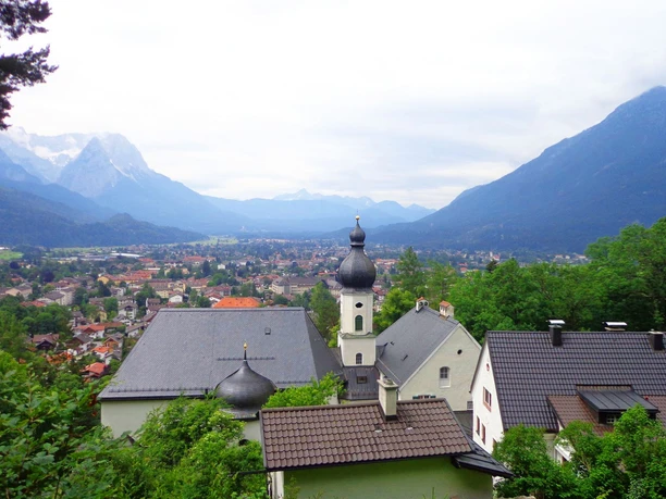 St. Anton Blick über Garmisch-Partenkirchen mit Zwiebelturm und umliegenden Bergen unter wolkigem Himmel
