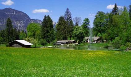 Kochelberg-Alm Grüne Wiese vor einem Teich mit Springbrunnen, umgeben von Hütten, Bäumen und Bergkulisse