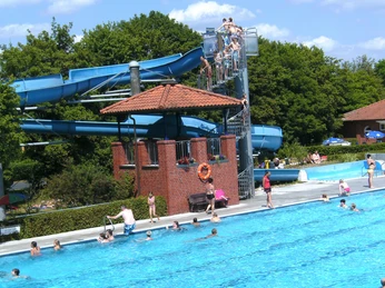 Freibad Hengstforde Das Bild zeigt ein belebtes Freibad mit vielen Menschen im und um das Wasser. Ein großer blauer Wasserpark mit rutschigen Röhren ragt links hervor, während Badegäste im klaren Becken schwimmen.