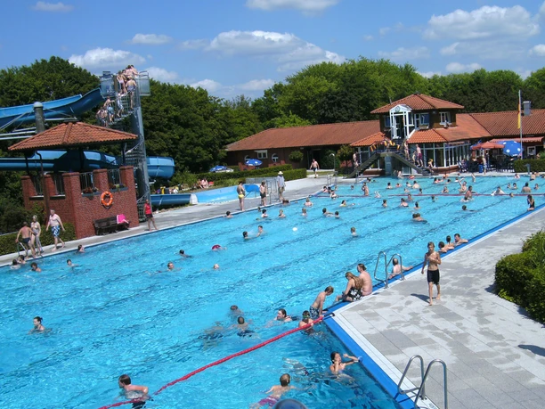 Schwimmvergnügen im Freibad Menschen genießen das Schwimmen in einem großen, sonnigen Freibad mit Rutschen und Liegewiese.