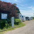 Wohnmobilstellplatz mit mehreren Fahrzeugen neben grünen Büschen und einer Informationstafel unter blauem Himmel.