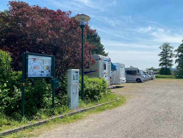 Wohnmobilstellplatz mit mehreren Fahrzeugen neben grünen Büschen und einer Informationstafel unter blauem Himmel.