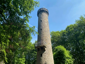 Steinerner Wilhelmsturm ragt vor blauem Himmel auf, umgeben von dichtem, grünem Wald im Sommer.