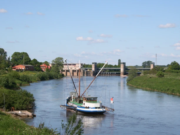 The Aalschocker Drakenburg on the Weser River in front of a lock, against a backdrop of blue skies and green riverbanks.