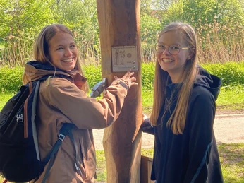 Hörstation Rheine 02-Hörstation Zwei junge Frauen lächeln, während sie aufmerksam eine interaktive Hörstation im Freien bedienen.