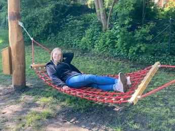 Hörstation 02 Rheine-Hängematte 2 Eine Frau entspannt sich lächelnd in einer roten Seilhängematte an einem sonnigen Tag, umgeben von Bäumen.