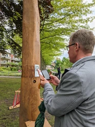 Hörtstation 02 Rheine-Bürgermeister Dr. Lüttmann Ein Mann scannt einen QR-Code an einer hölzernen Hörstation in einem parkähnlichen Umfeld.