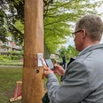 Hörtstation 02 Rheine-Bürgermeister Dr. Lüttmann Ein Mann scannt einen QR-Code an einer hölzernen Hörstation in einem parkähnlichen Umfeld.