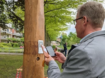 Hörtstation 02 Rheine-Bürgermeister Dr. Lüttmann Ein Mann scannt einen QR-Code an einer hölzernen Hörstation in einem parkähnlichen Umfeld.