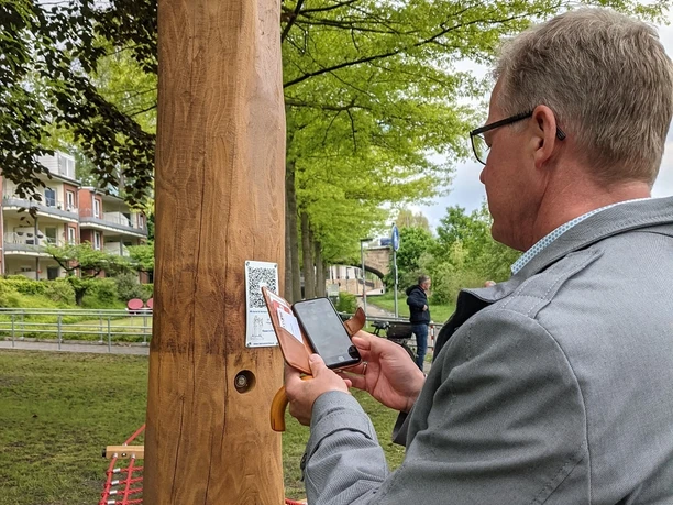 Hörtstation 02 Rheine-Bürgermeister Dr. Lüttmann Ein Mann scannt einen QR-Code an einer hölzernen Hörstation in einem parkähnlichen Umfeld.
