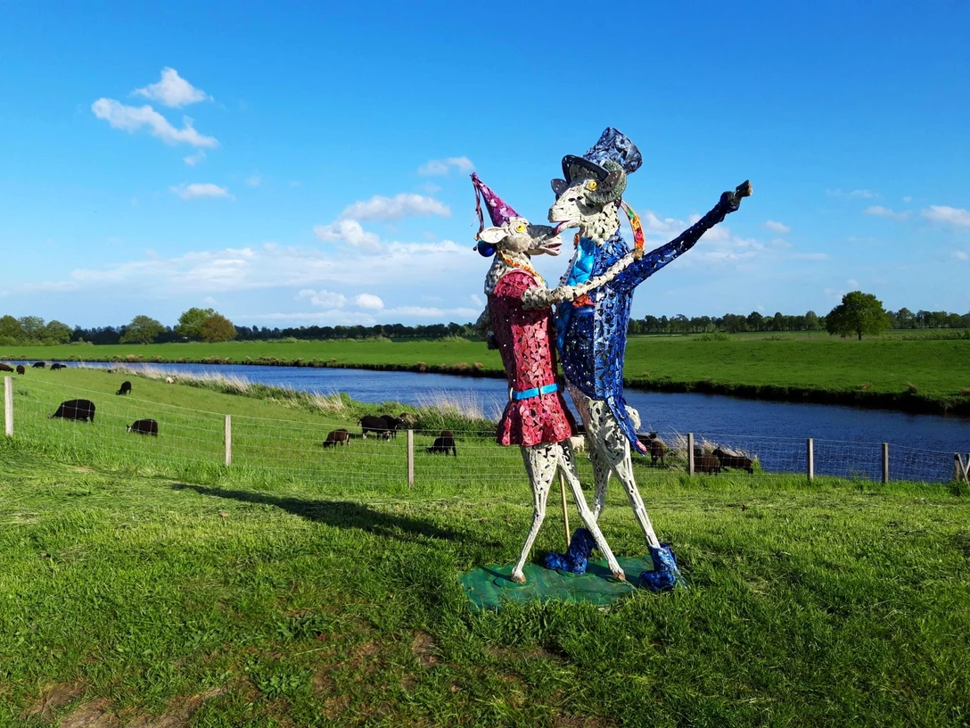 Tanz auf dem Deich Eine bunte Skulptur von zwei tanzenden Figuren steht auf einer grünen Wiese, flankiert von einem Fluss und Kühen im Hintergrund.