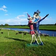 Tanz auf dem Deich Eine bunte Skulptur von zwei tanzenden Figuren steht auf einer grünen Wiese, flankiert von einem Fluss und Kühen im Hintergrund.
