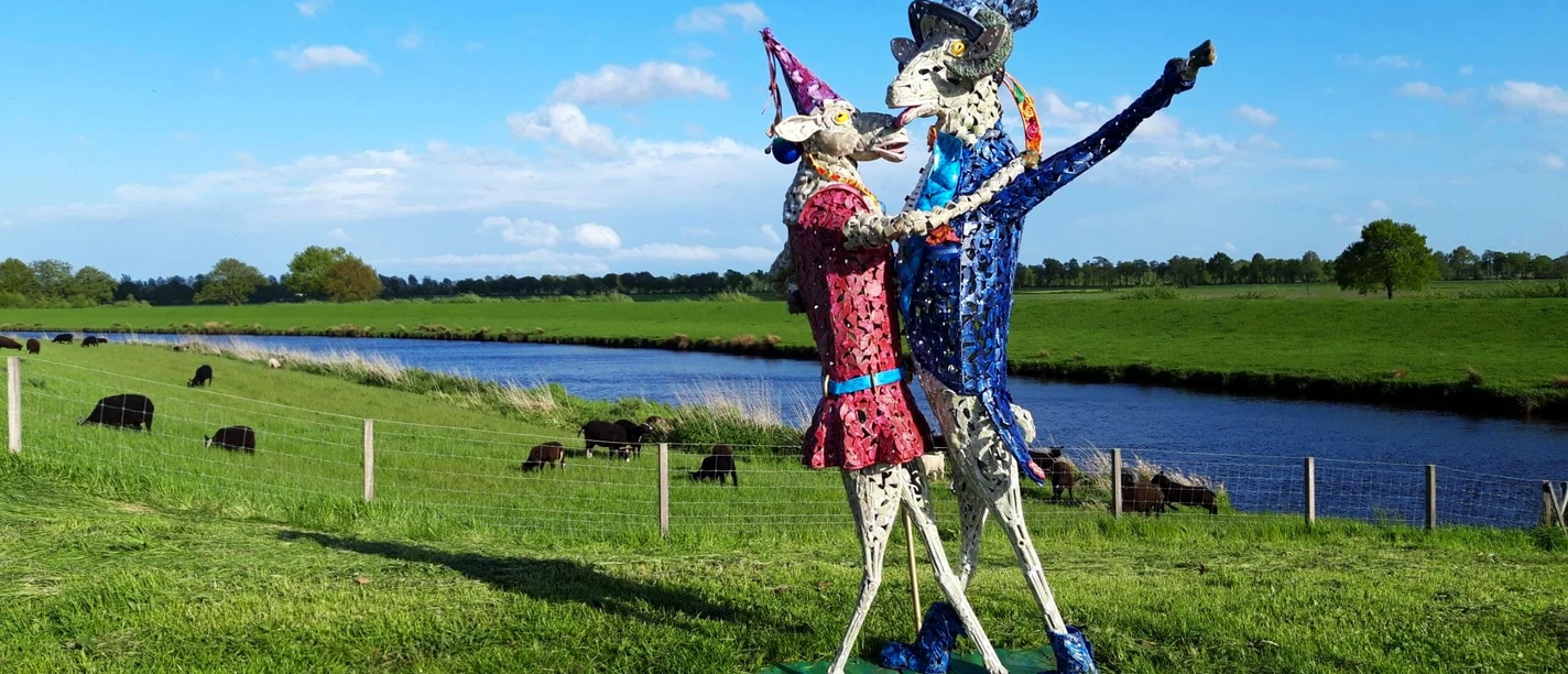 Tanz auf dem Deich Eine bunte Skulptur von zwei tanzenden Figuren steht auf einer grünen Wiese, flankiert von einem Fluss und Kühen im Hintergrund.