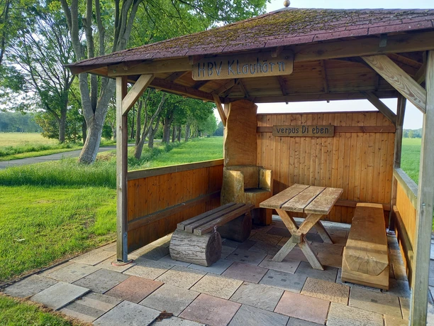 Rastplatz in Klauhörn Der Rastplatz in Klauhörn zeigt einen überdachten Unterstand mit Holztisch und Bänken auf gepflastertem Boden. Im Hintergrund sind grüne Wiesen und hohe Bäume zu sehen, die eine malerische Landschaft bilden.