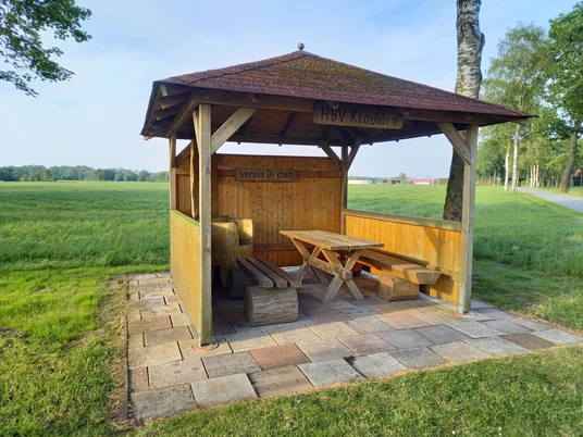Erholsame Pause in Klauhörn Ein hölzerner Pavillon mit Sitzgelegenheiten steht auf einer gepflasterten Fläche inmitten einer grünen Wiese, ideal für eine erholsame Pause.