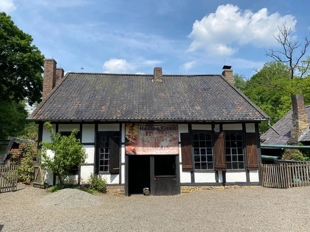 Oelchenshammer Traditionelles Fachwerkhaus mit grauem Dach und Schornsteinen, umgeben von Bäumen, bei sonnigem Wetter.