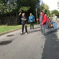 Boßelspaß Auf einer ruhigen Landstraße wird das traditionelle Boßeln gespielt. Eine Gruppe von Menschen, darunter eine Frau im Vordergrund, konzentriert sich auf das Werfen einer roten Boßelkugel über die Asphaltstraße, begleitet von lächelnden Teilnehmern und einem Bollerwagen voller Zubehör im Hintergrund. Die umgebende grüne Landschaft schafft eine friedliche Kulisse an diesem sonnigen Tag.