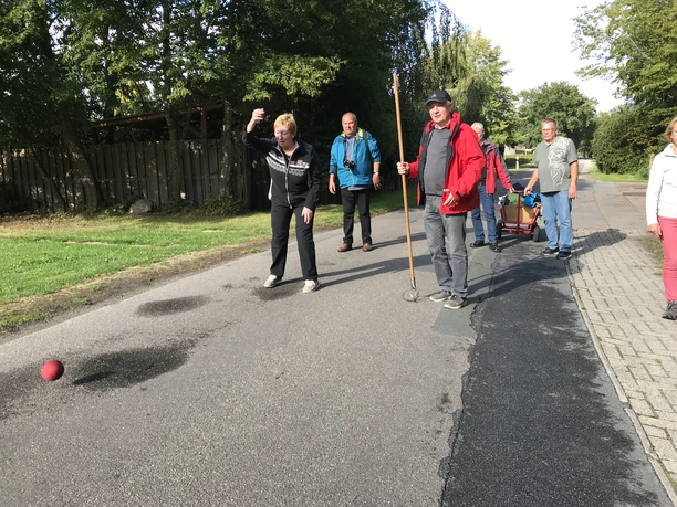Boßelspaß Auf einer ruhigen Landstraße wird das traditionelle Boßeln gespielt. Eine Gruppe von Menschen, darunter eine Frau im Vordergrund, konzentriert sich auf das Werfen einer roten Boßelkugel über die Asphaltstraße, begleitet von lächelnden Teilnehmern und einem Bollerwagen voller Zubehör im Hintergrund. Die umgebende grüne Landschaft schafft eine friedliche Kulisse an diesem sonnigen Tag.