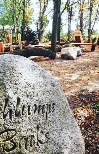Stein mit Gravur "Schlumps Bracks" in einem Waldspielplatz mit Holzklettergeräten und Bänken.