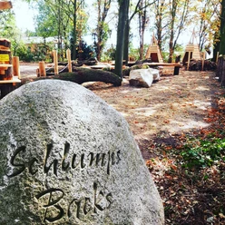 Schlumps Backs.jpg Stein mit Gravur "Schlumps Bracks" in einem Waldspielplatz mit Holzklettergeräten und Bänken.