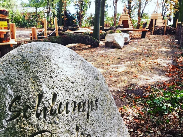 Schlumps Backs.jpg Stein mit Gravur "Schlumps Bracks" in einem Waldspielplatz mit Holzklettergeräten und Bänken.