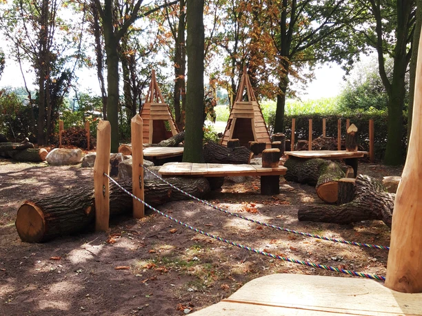 Holzspielplatz im Wald mit großen Baumstämmen, Seilen zum Balancieren und hölzernen Schutzhütten.