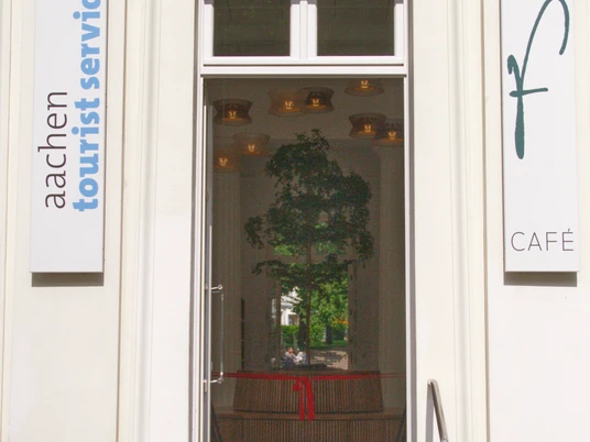 Eingang zur Touristeninformation im Elisenbrunnen. Rechts an der Wand hängt ein Schild mit einem großen "f" und "Café" und links ein Schild mit dem Aufdruck "aachen tourist service". Im Inneren hängt ein Band mit roter Schleife und ein kleiner Baum, der von einer runden Sitzbank umrandet wird.