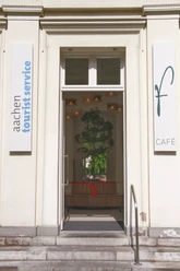 Neueröffnung TI Außenansicht Eingang zur Touristeninformation im Elisenbrunnen. Rechts an der Wand hängt ein Schild mit einem großen "f" und "Café" und links ein Schild mit dem Aufdruck "aachen tourist service". Im Inneren hängt ein Band mit roter Schleife und ein kleiner Baum, der von einer runden Sitzbank umrandet wird.
