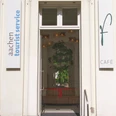 Neueröffnung TI Außenansicht Eingang zur Touristeninformation im Elisenbrunnen. Rechts an der Wand hängt ein Schild mit einem großen "f" und "Café" und links ein Schild mit dem Aufdruck "aachen tourist service". Im Inneren hängt ein Band mit roter Schleife und ein kleiner Baum, der von einer runden Sitzbank umrandet wird.