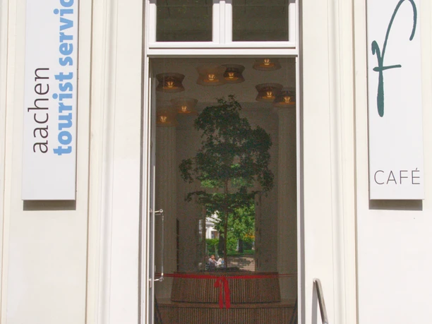 Neueröffnung TI Außenansicht Eingang zur Touristeninformation im Elisenbrunnen. Rechts an der Wand hängt ein Schild mit einem großen "f" und "Café" und links ein Schild mit dem Aufdruck "aachen tourist service". Im Inneren hängt ein Band mit roter Schleife und ein kleiner Baum, der von einer runden Sitzbank umrandet wird.