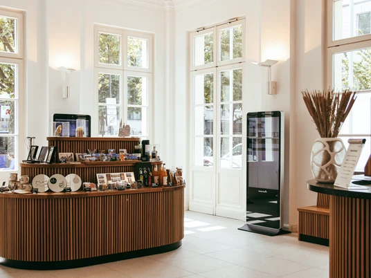 Offener, heller Raum mit hohen Fenstern und einer Auswahl an Produkten und Souvenirs auf einem ovalen Tisch. Rechts an der Wand steht eine digitale, interaktive Stele mit großem Bildschirm.