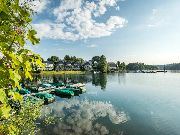 Bevertalsperre Ein idyllischer See mit Booten im Vordergrund und malerischen Häusern am Ufer, spiegelnde Wasserfläche.