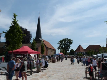 troedelmarkt-rehburg-stadtplatz-staende.JPG Ein Marktplatz mit Ständen, Menschen bummeln; im Hintergrund eine Kirche und Bäume sichtbar.