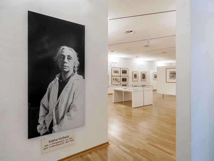 Käthe Kollwitz Museum Cologne Das Bild zeigt einen hellen Raum im Käthe Kollwitz Museum in Köln. Links ist ein großes Schwarz-Weiß-Porträt von Käthe Kollwitz in einem weißen Kittel vor einer Wand zu sehen. Im Hintergrund befinden sich Ausstellungsstücke in Vitrinen und an Wänden auf einem Parkettboden.The picture shows a bright room in the Käthe Kollwitz Museum in Cologne. On the left is a large black and white portrait of Käthe Kollwitz in a white coat in front of a wall. In the background are exhibits in display cases and on walls on a parquet floor.