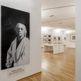 Käthe Kollwitz Museum Köln Das Bild zeigt einen hellen Raum im Käthe Kollwitz Museum in Köln. Links ist ein großes Schwarz-Weiß-Porträt von Käthe Kollwitz in einem weißen Kittel vor einer Wand zu sehen. Im Hintergrund befinden sich Ausstellungsstücke in Vitrinen und an Wänden auf einem Parkettboden.The picture shows a bright room in the Käthe Kollwitz Museum in Cologne. On the left is a large black and white portrait of Käthe Kollwitz in a white coat in front of a wall. In the background are exhibits in display cases and on walls on a parquet floor.