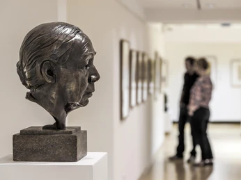 Käthe Kollwitz Museum Köln Eine Bronzebüste der Künstlerin Käthe Kollwitz steht seitlich auf einem Podest, während im Hintergrund zwei Personen in einer Galerie die Kunstwerke betrachten.A bronze bust of a woman's profile is displayed in the foreground, with visitors observing artworks in a lit hallway in the background at the Käthe Kollwitz Museum in Cologne.