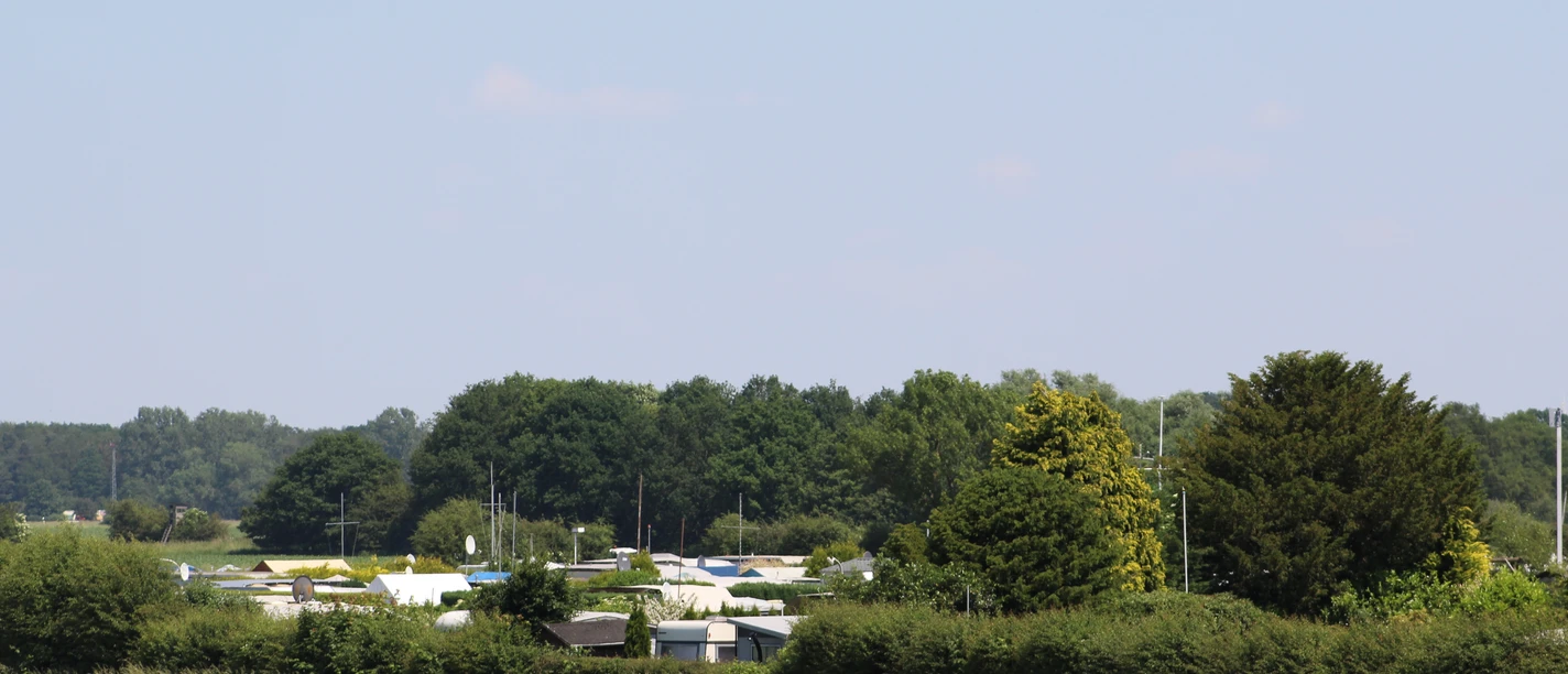 Camping met caravan tussen groene struiken en bomen, bos in de verte op de achtergrond.