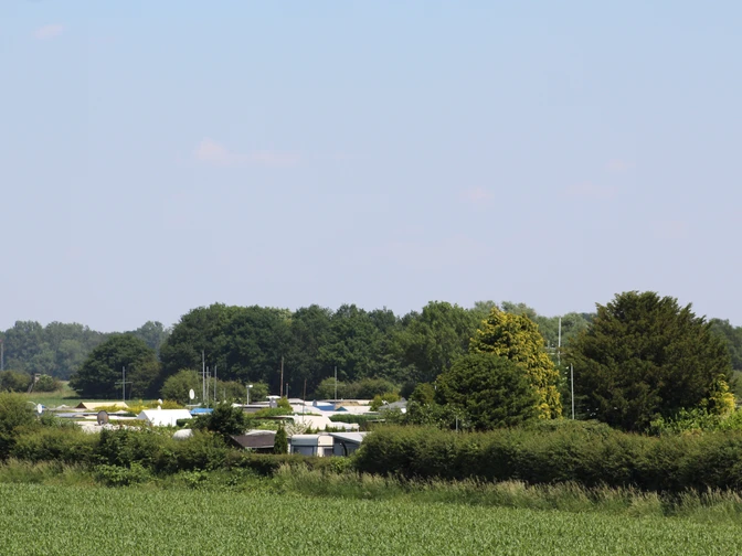 Campingplatz mit Wohnwagen zwischen grünen Büschen und Bäumen, entferntes Waldstück im Hintergrund.