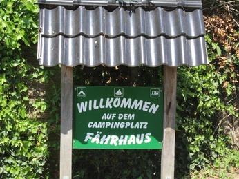 Grünes Schild mit weißer Aufschrift "Willkommen auf dem Campingplatz Fährhaus", umrahmt von Pflanzen.Green sign with white lettering "Welcome to the Fährhaus campsite", surrounded by plants.Grønt skilt med hvid skrift "Velkommen til campingpladsen Fährhaus", indrammet af planter.Groen bord met witte letters "Welkom op camping Fährhaus", omlijst door planten.