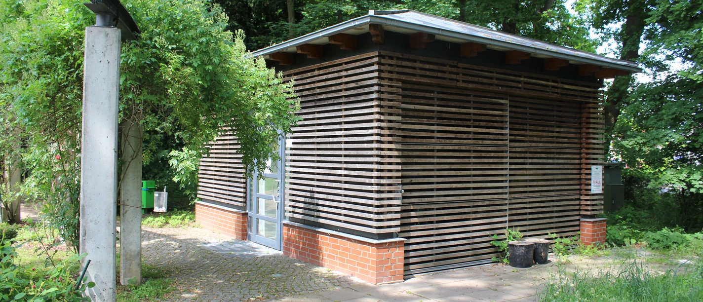 Öffentliches Toilettenhäuschen im Schlosspark Etelsen mit natürlicher Holzverkleidung, umgeben von Grün.