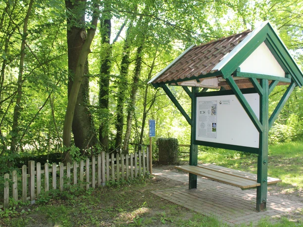 Kneippanlage mit hölzernem Unterstand und Sitzbank, umgeben von dichten, grünen Bäumen und einem kleinen Zaun.