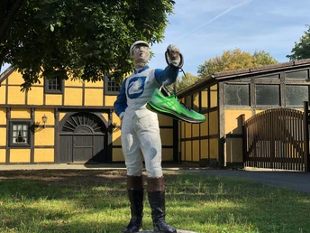 Figur eines Mannes in Reitkleidung präsentiert einen grünen Schuh auf einem Anwesen im Fachwerkstil.