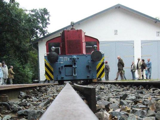 Historische Lokomotive auf Schienen, umgeben von Besuchern vor einem weiß gestrichenen Gebäude.