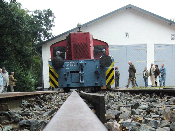 Historische Lokomotive auf Schienen, umgeben von Besuchern vor einem weiß gestrichenen Gebäude.