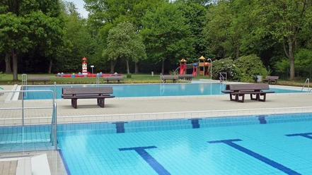 Freibad mit blauem Becken und Sprungturm, umgeben von üppigen Bäumen und bunter Kinderspielanlage.
