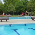 Freibad mit blauem Becken und Sprungturm, umgeben von üppigen Bäumen und bunter Kinderspielanlage.
